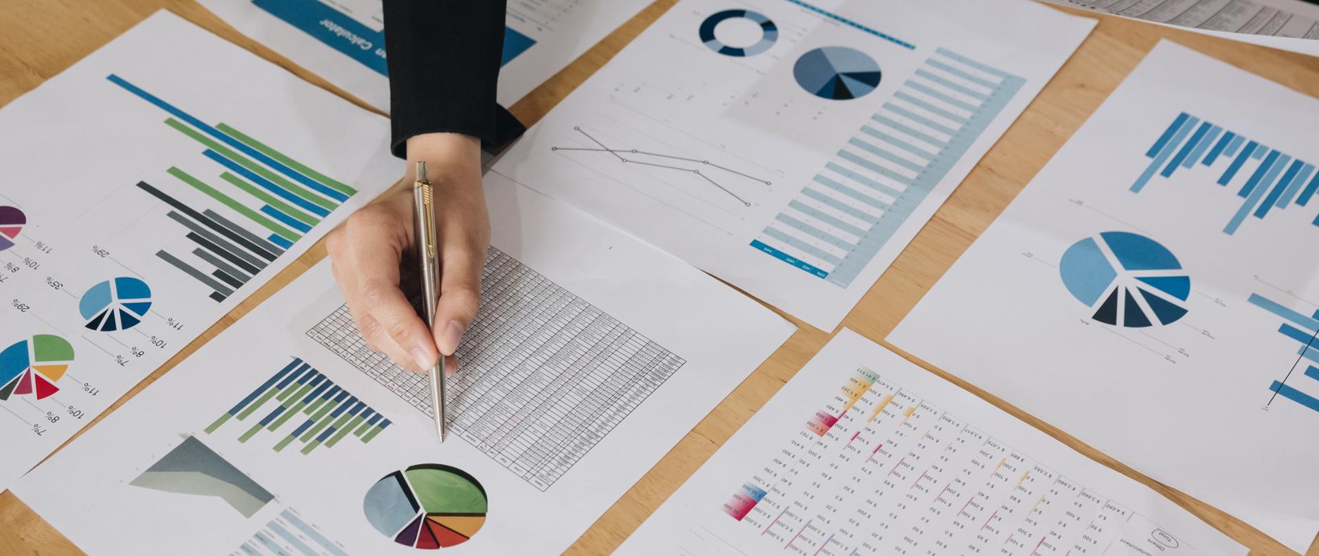 Na zdjęciu osoba trzymająca długopis, przed nią dokumenty z wykresami.