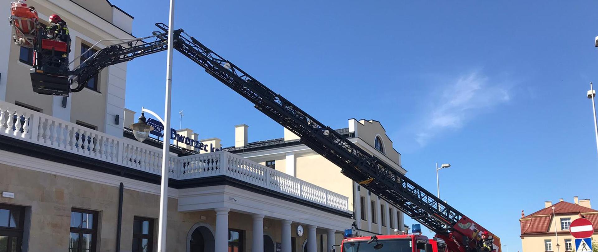 Na zdjęciu widać na pierwszym planie strażacki samochód typu drabina. Samochód jest w kolorze czerwonym. Obok pojazdu przechodzi ratownik ubrany w czarne ubranie bojowe. Na głowie ma czerwony hełm. Samochód stoi przed budynkiem dworca kolejowego z którego dachu przy użyciu drabiny mechanicznej ewakuują poszkodowaną osobę. Osoba ewakuowana jest na desce ortopedycznej umocowanej do kosza mechanicznej drabiny. W koszu znajduje się strażak ubrany w czarne ubranie bojowe. Na głowie ma czerwony hełm. Strażak asekuruje poszkodowaną osobę przy ewakuacji. W tle widoczne są zabudowania.