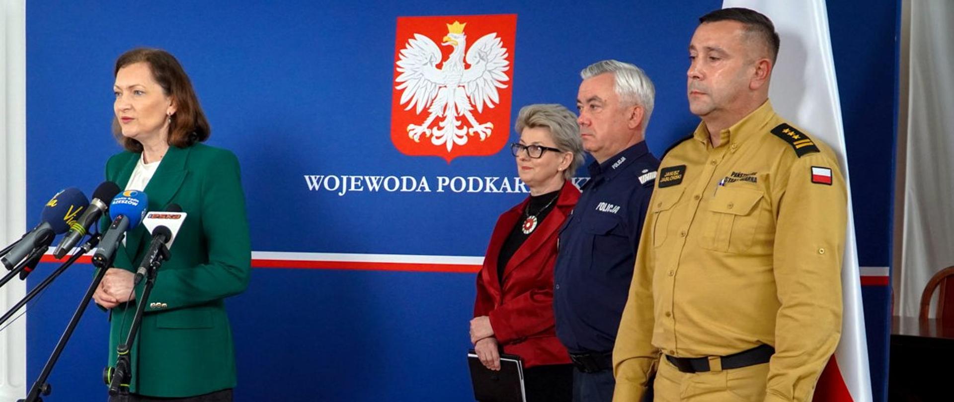 Zdjęcie wykonane w pomieszczeniu, podczas konferencji prasowej. Przed mikrofonami stoi wojewoda podkarpacki. Po prawej stronie stoi trzech uczestników konferencji, wśród nich oficer państwowej straży pożarnej w stopniu starszego brygadiera oraz oficer Policji w stopniu nadinspektora. Uczestnicy konferencji stoją na tle ścianki na której umieszczone jest godło RP oraz napis wojewoda podkarpacki. 