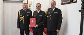 Trzech funkcjonariuszy Państwowej Straży Pożarnej w mundurach wyjściowych ze sznurem stoi obok siebie strażak będący w środku trzyma czerwoną teczkę ze złotym orzełkiem i napisem za nimi stoją dwie flagi Polski wisi obraz oraz znajdują się drzwi. 