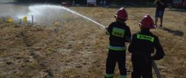 Zawody sportowo-pożarnicze w Jeziorach. Plac ćwiczeń. Trwa jedna z konkurencji zawodów - ćwiczenie bojowe. Końcowy etap ćwiczenia-strącanie przy pomocy strumienia wody, pachołków ustawionych na postumentach. Zadania te wykonuje jedna rota - 2 strażaków, członków MDP, którzy mają czerwone hełmy. W tle drzewa i przeszkody.