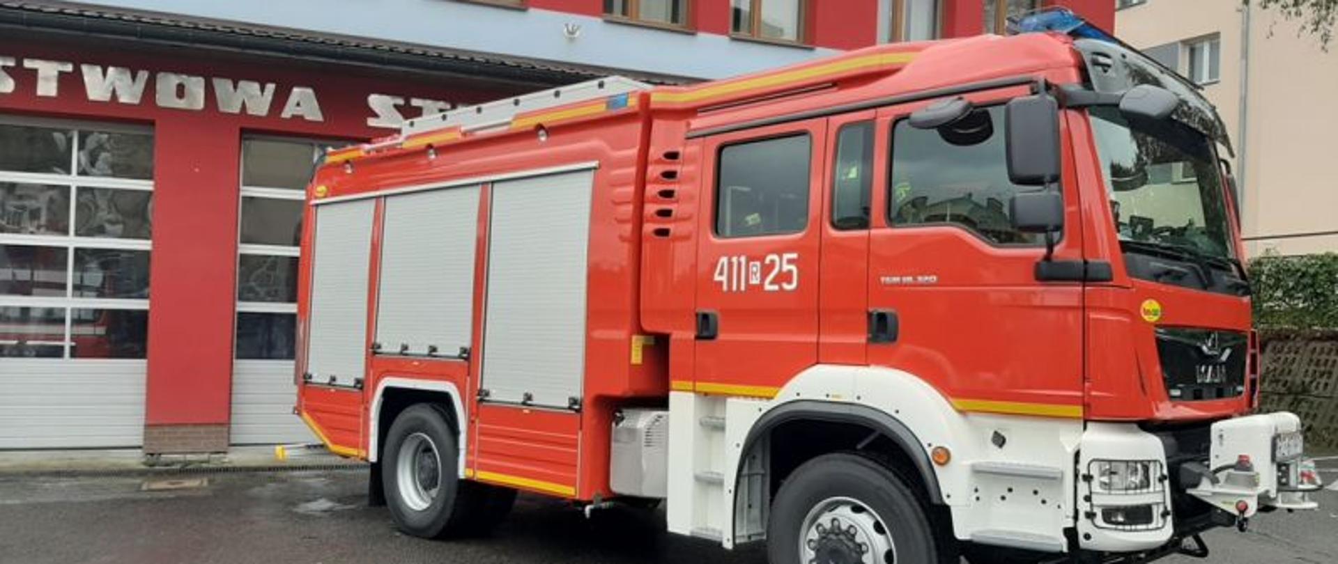 Fotografia przedstawia czerwony, nowy, ciężki samochód ratowniczo-gaśniczy na tle budynku remizy Komendy Powiatowej Państwowej Straży Pożarnej w Brzozowie. Na fotografii widać bojowy samochód pożarniczy, jego przód i prawy bok. 