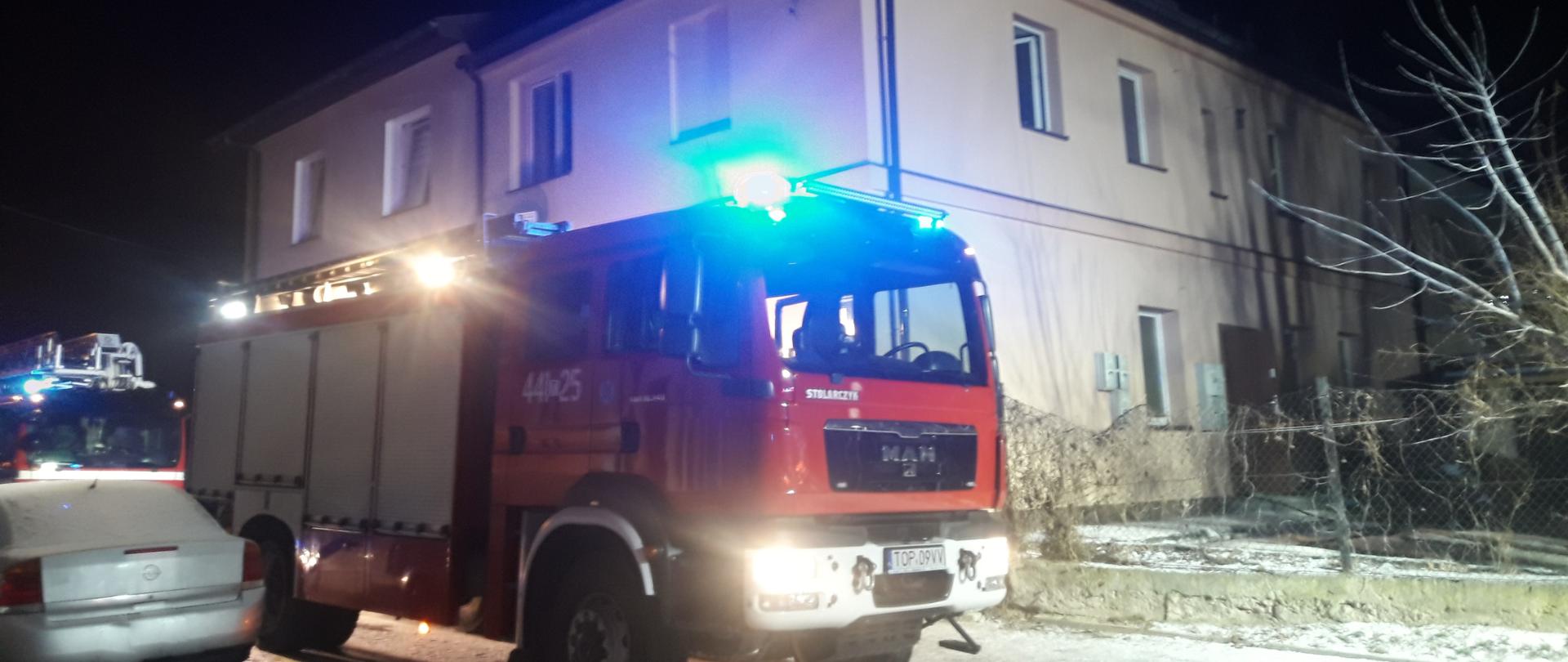 Zdjęcie przedstawia dwa czerwone samochody straży pożarnej oraz samochód osobowy. W tle samochodów widać budynek mieszkalny wielorodzinny