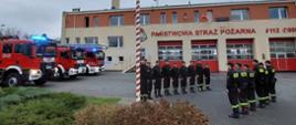 Zdjęcie przedstawia strażaków podczas uroczystego podnoszenia flagi na maszt, w tle widać samochody strażackie oraz budynek Komendy Powiatowej PSP w Pruszczu Gdańskim.