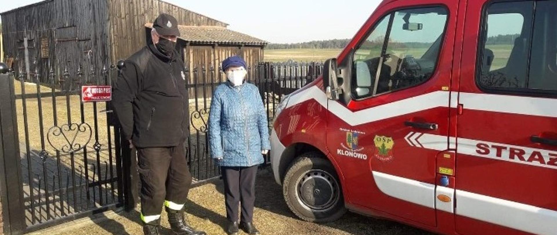 Pierwszy plan: druh z Ochotniczej Straży Pożarnej w Klonowie oraz kobieta w podeszłym wieku stoją obok pojazdu strażackiego.
Drugi plan: budynki gospodarcze.