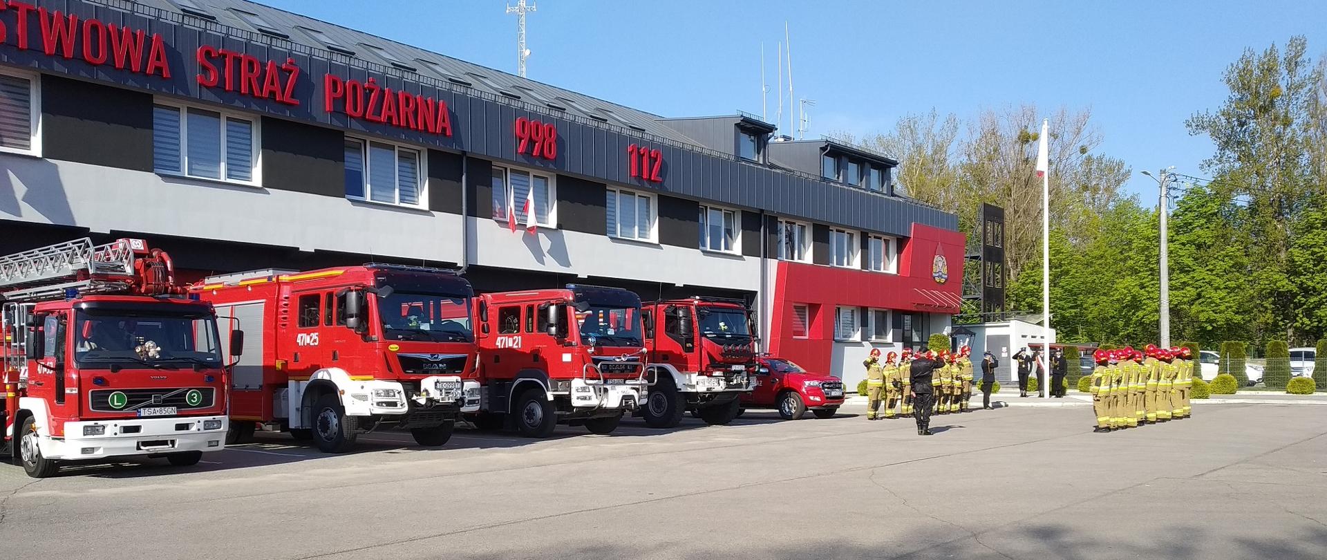 Zdjęcie zrobione w dzień. Przed remizą strażacką stoją stoi pięć samochodów strażackich. Przed nimi strażacy w dwóch szeregach w ubraniach bojowych. W tle widać maszt, na którym powiewa biało-czerwona flaga. Pod masztem stoi trzech strażaków w mundurach reprezentacyjnych. Strażacy ci salutują.