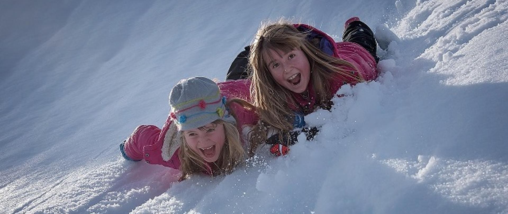 Dwie uśmiechnięte dziewczynki leżące na brzuchu w zimowych ubraniach na górce pełnej śniegu