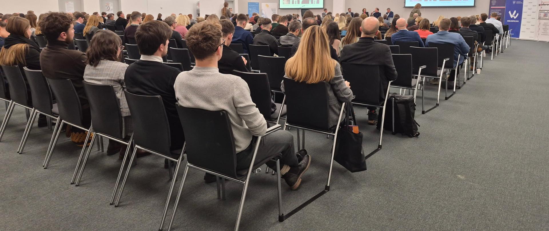 Fotografia przedstawia salę konferencyjną. Na pierwszym planie siedzą uczestnicy spotkania na krzesłach, odwróceni tyłem do zdjęcia. W oddali widać na czterech projektorach wyświetloną nazwę panelu wraz z panelistami.