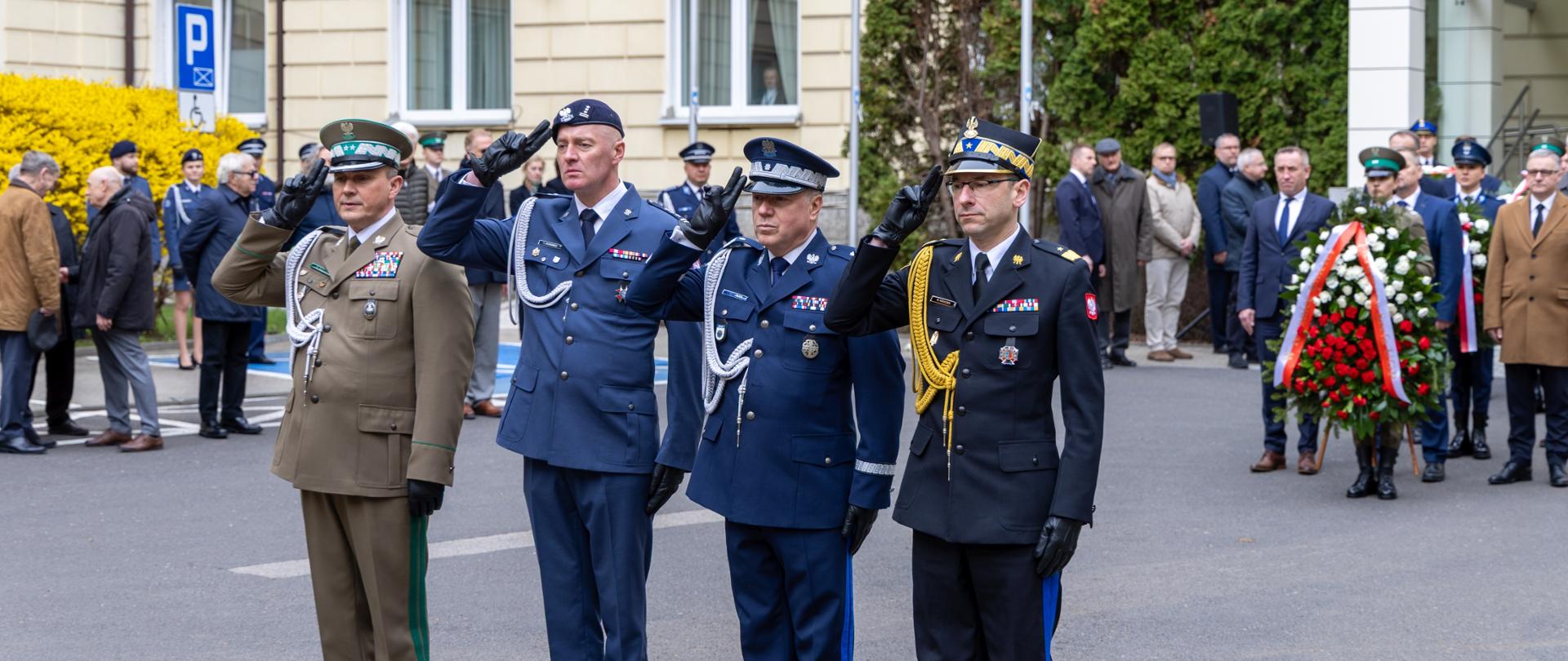 Grupa oficerów służb mundurowych oddaje salut podczas ceremonii, w tle uczestnicy uroczystości i złożone wieńce.
