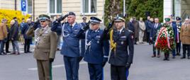 Grupa oficerów służb mundurowych oddaje salut podczas ceremonii, w tle uczestnicy uroczystości i złożone wieńce.
