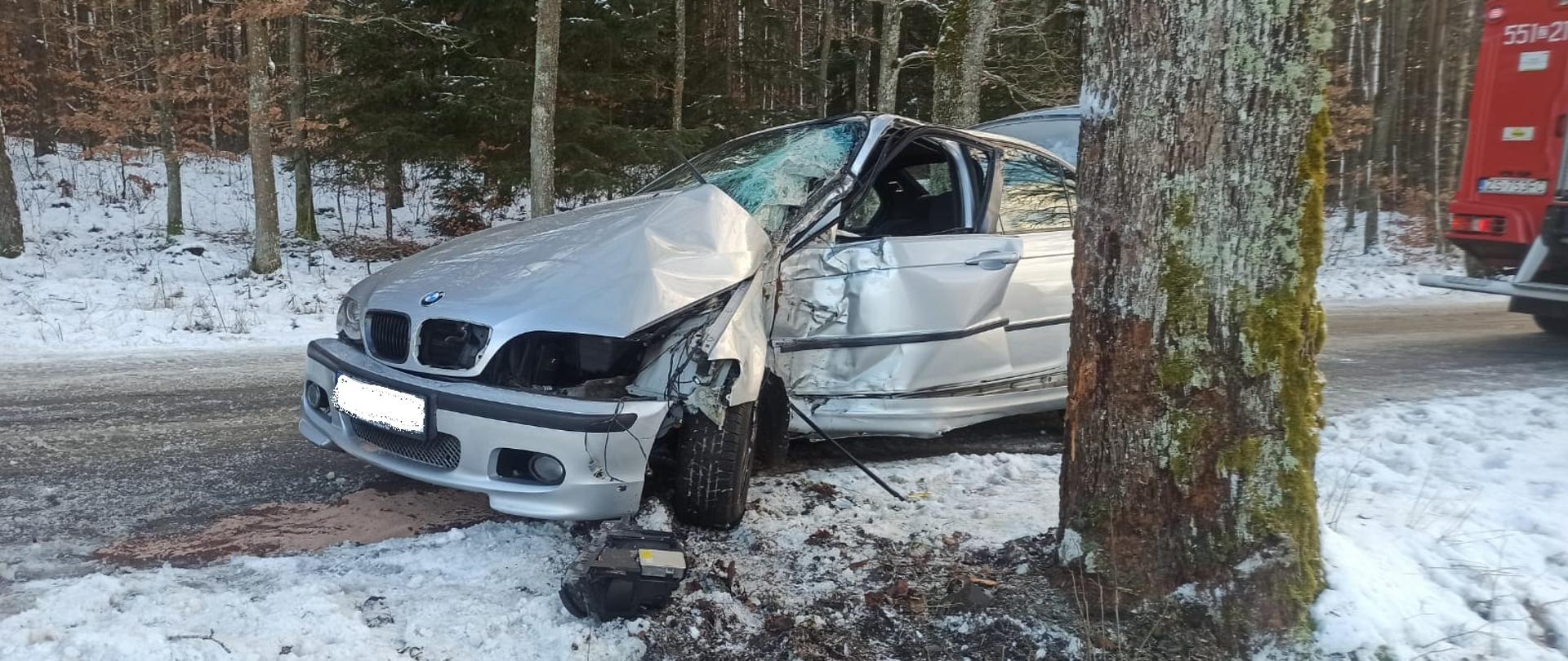 Zdjęcie przedstawia drogę pokrytą błotem pośniegowym na której znajduję się srebrne auto BMW po uderzeniu bocznym w drzewo. Przód auta jest uszkodzony, maska wygięta do góry, drzwi boczne kierowcy mocno wgniecione do środka a szyba pobita. Z pod pojazdu wyciekają płyny eksploatacyjne. W tle widać pole i drzewa. Na pierwszym planie widać drzewo w które uderzyło auto a po prawej stronie widać wóz strażacki.