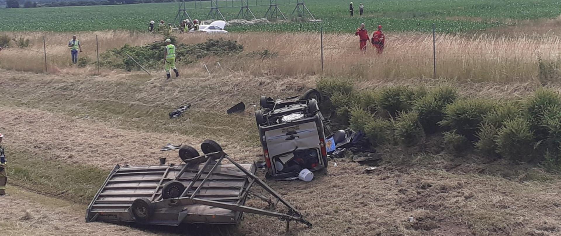 Dzień. Na pierwszym planie bus oraz przyczepa leżące na dachu na poboczu autostrady. Na drugim planie widoczny biały samochód osobowy poza ogrodzeniem autostrady. Strażacy i ratownicy medyczni prowadzą działania.