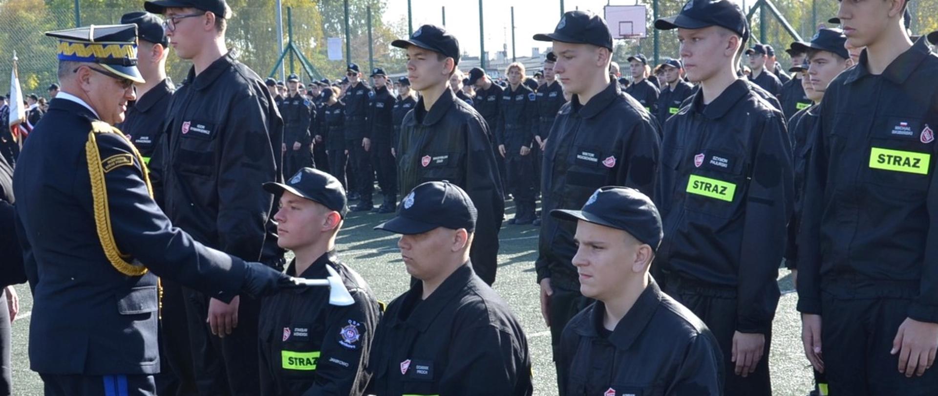 Komendant PSP , generał pasuje ucznia, kładzie mu na prawe ramię strażacki toporek. Uczeń pasowany klęczy na prawe kolano. Za nimi kolejni.