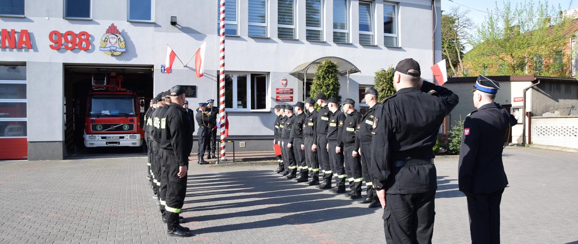 Uroczysta zmiana służby przed budynkiem KP PSP w Mińsku Mazowieckim. Poczet flagowy przygotowuje się do wciągnięcia flagi na maszt. Dwie zmiany służbowe stoją w szeregach na przeciwko siebie. Wszyscy stoją na baczność