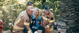 Trzech strażaków PSP niesie rannego kobietę z unieruchomioną nogą. Poszkodowana ubrana w niebieską bluzę i czarne spodnie trzymana jest w pozycji siedzącej. W tle widać innych ratowników i miejsce zdarzenia w lesie.