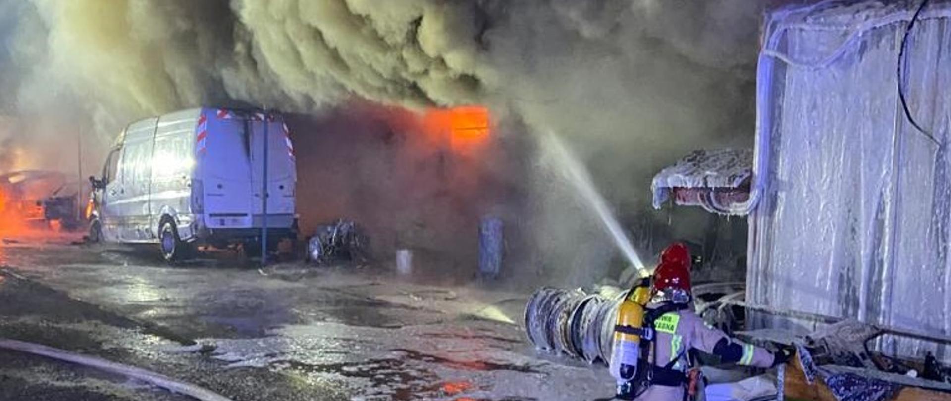 Zdjęcie przedstawia rozwinięty pożar warsztatu samochodowego. Po lewej stronie stoi zniszczony przez ogień samochód dostawczy. W głębi widać palący się ogień oraz kłęby gęstego dymu. Po prawej stronie dwóch ratowników podaje wodę zza przeszkody terenowej. 