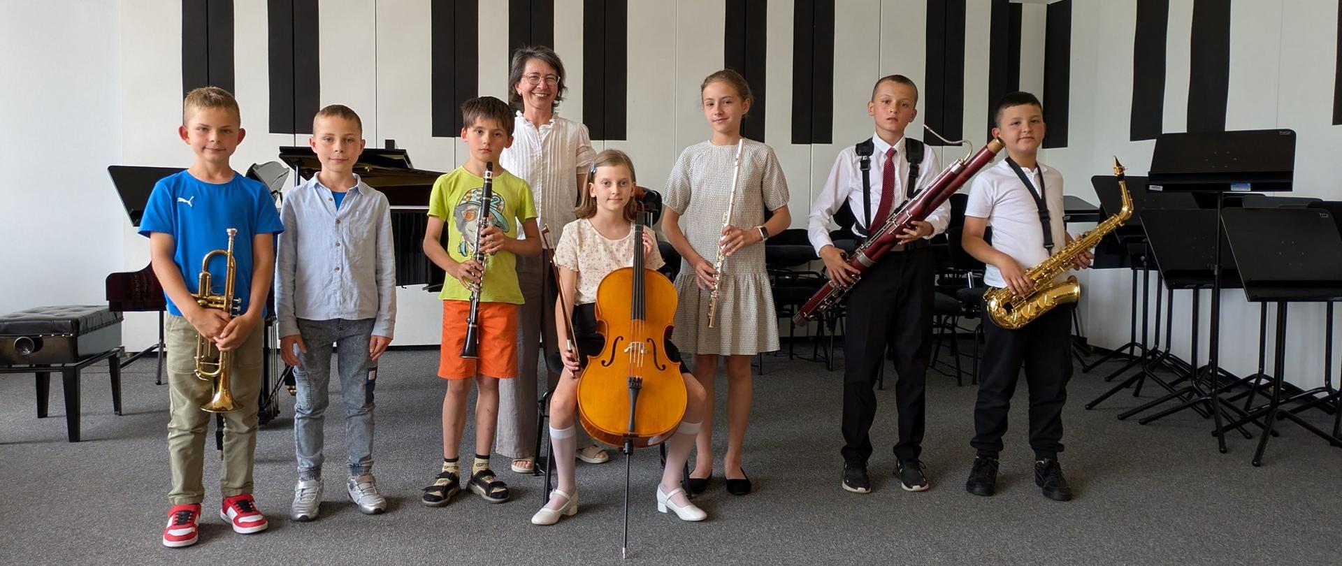 Wszyscy wykonawcy stoją ze swoimi instrumentami na scenie, za nimi stoi nauczycielka.