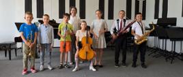 Wszyscy wykonawcy stoją ze swoimi instrumentami na scenie, za nimi stoi nauczycielka.