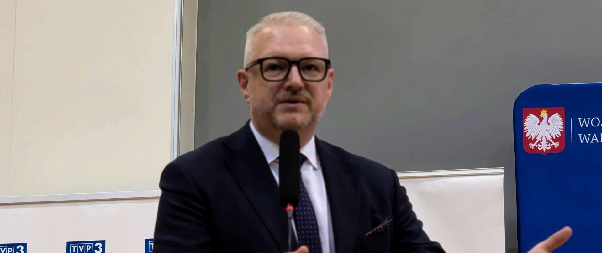 Mężczyzna w granatowym garniturze, wojewoda, przemawia na mównicy, gestykuluje