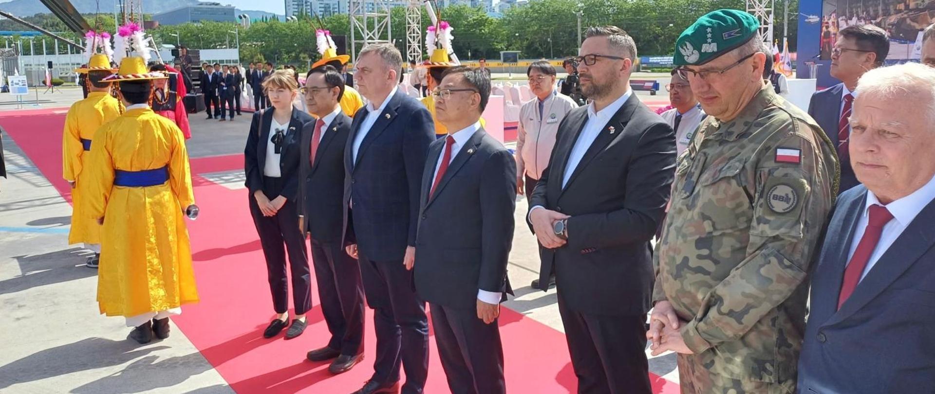 Na zdjęciu w szeregu, na czerwonym dywanie stoi siedem osób. Od lewej strony idą dwie osoby w żółtych kimonach przepasane niebieskim paskiem. 