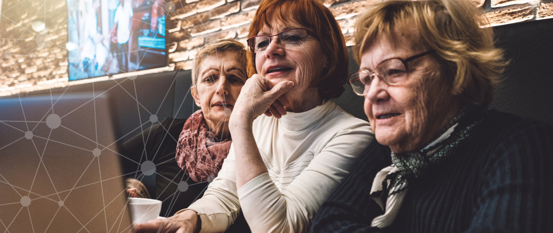 Trzy seniorki siedzą w kawiarni i patrzą wspólnie na ekran laptopa.