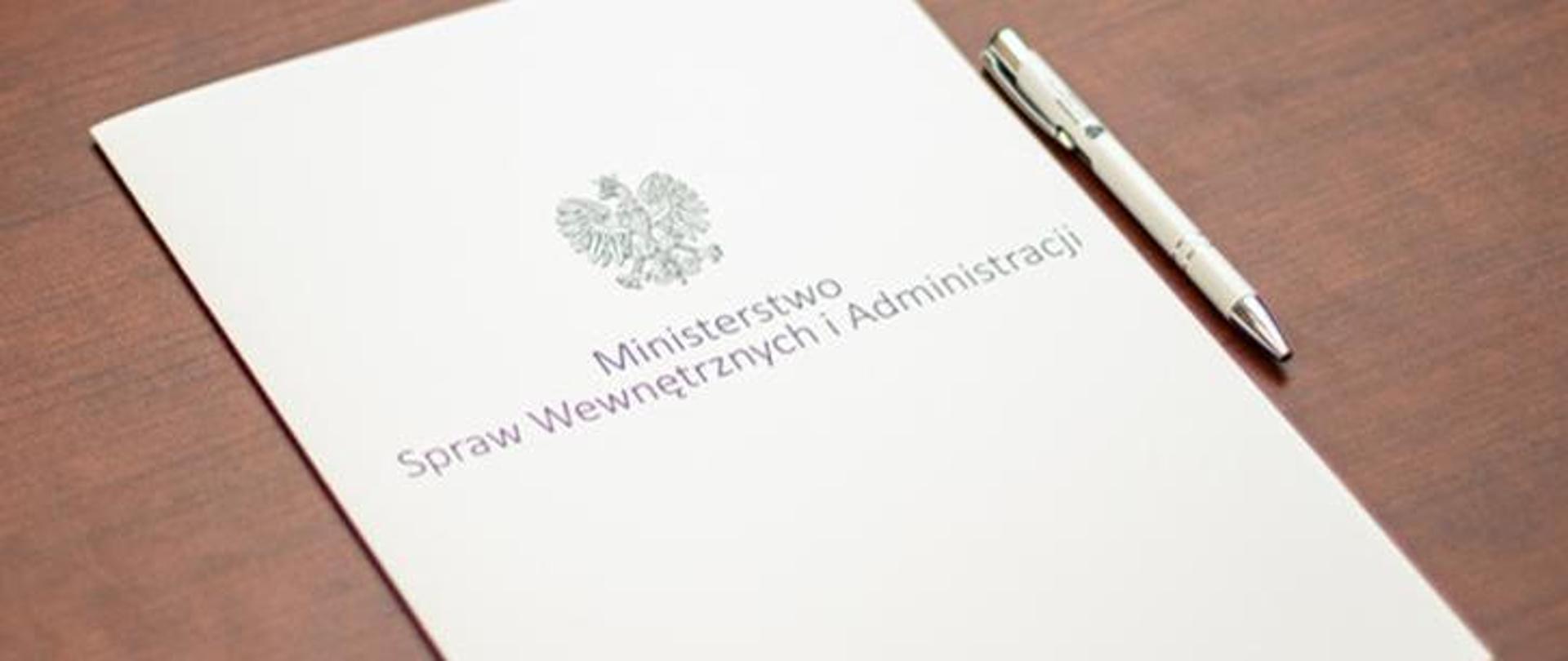 Zdjęcie przedstawia teczkę Ministerstwa Spraw Wewnętrznych i Administracji oraz długopis, które leżą na biurku