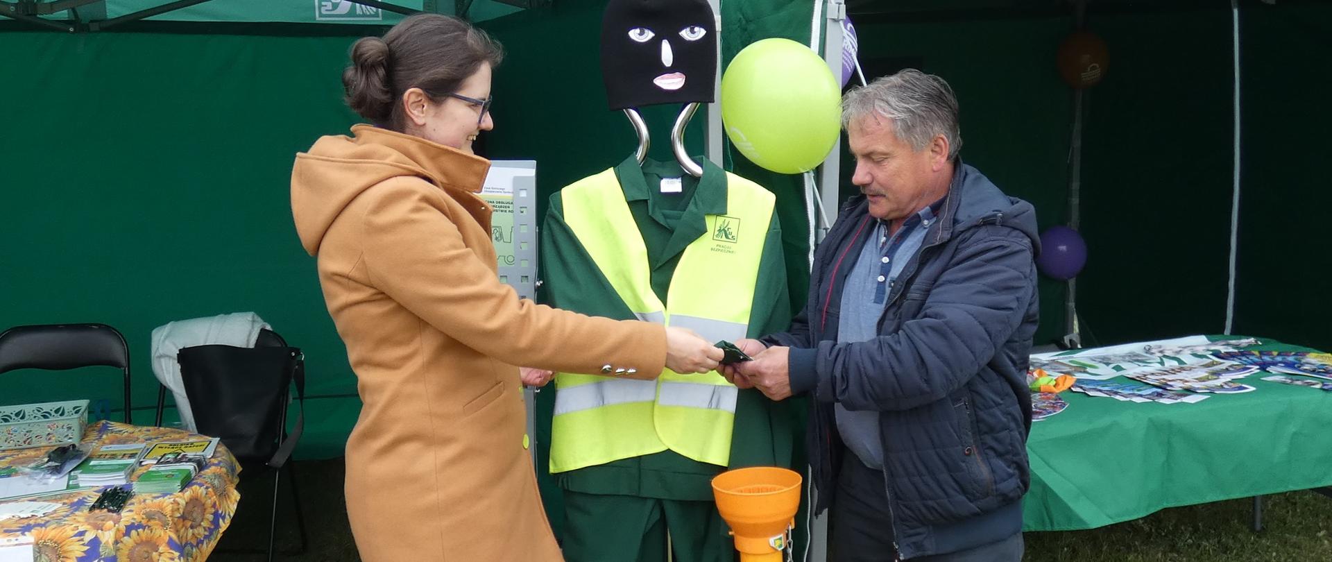 Mężczyzna i kobieta stoją obok manekina ubranego w odzież roboczą z elementami odblaskowymi. W tle widoczne zielone namioty z logo KRUS i ARiMR oraz stoły z materiałami informacyjnymi. 