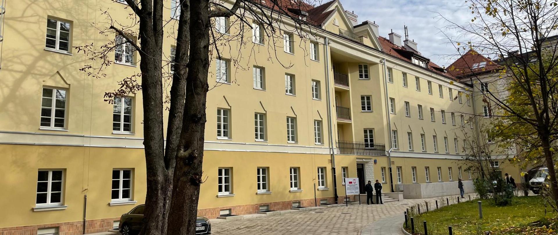 Zdjęcie na dworze, widok pod kątem na ścianę żółtego czteropiętrowego budynku ze skośnym krytym czerwoną dachówką dachem, przed budynkiem podjazd z szarej trylinki.