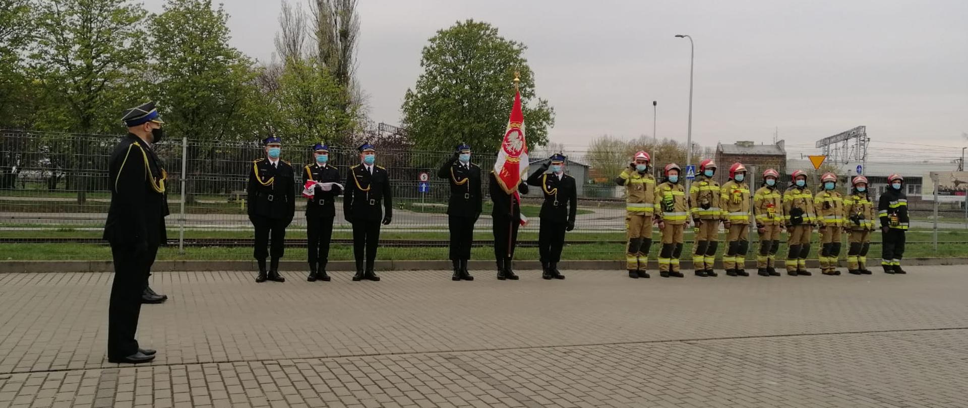 Zdjęcie przedstawia uroczyste podniesienie flagi państwowej w Komendzie Powiatowej Państwowej Straży Pożarnej w Pruszkowie z okazji dnia Flagi. Na zdjęciu widoczny jest dowódca uroczystości, poczet flagowy i sztandarowy oraz zmiana służbowa JRG. 
