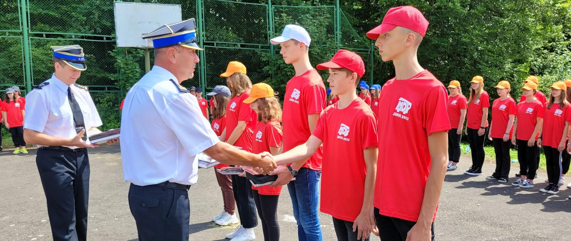 Zdjęcie zrobione na zewnątrz podczas uroczystej zbiórki Młodzieżowych Drużyn Pożarniczych. Oficer Państwowej Straży Pożarnej podaje dłoń chłopcu z Młodzieżowej Drużyny Pożarniczej. Obok niego stoi oficer Państwowej Straży Pożarnej. W tle widać uczestników obozu zebranych na uroczystej zbiórce.