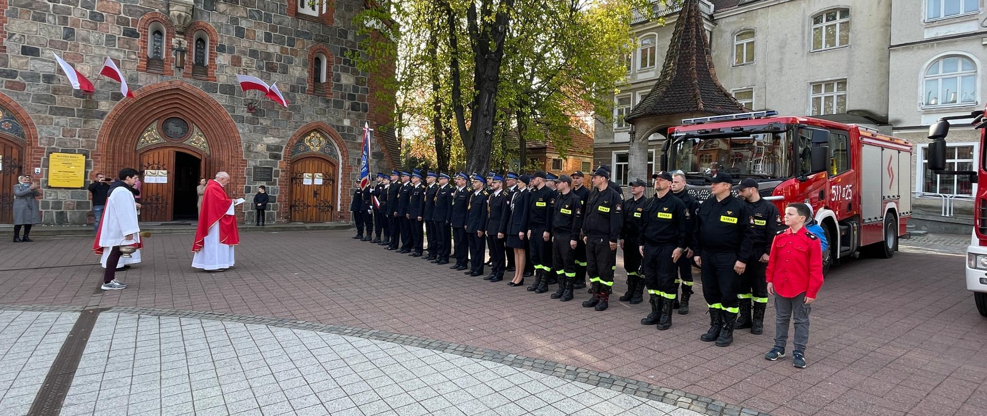 Sopoccy strażacy stoją w dwuszeregu na placu przed kościołem. Za nimi są samochody strażackie. Przed nimi stoi ksiądz i odmawia modlitwę. Część strażaków jest w mundurach galowych a część w koszarowych . Na końcu stoi członek Młodzieżowej Drużyny Pożarniczej OSP Sopot