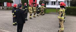 Zdjęcie przedstawia zmianę służbową strażaków na placu apelowym. Na czele szeregu na pierwszym planie stoi oficer ubrany w mundur wyjściowy z czapką rogatywką na głowie. Strażacy stoją frontem do siebie w szeregu, dziewięciu na lewej stronie i ośmiu po prawej stronie. Trzech strażaków po lewo z końca szeregu maszeruje krokiem defiladowym w kierunku masztu, środkowy trzyma w dłoniach flagę państwową, pozostali mają ręce wzdłuż ciała. Wszyscy strażacy są ubrani w ubrania bojowe typu Moratex, na głowie maja heł, na twarzy maseczki ochronne, na nogach kalosze. Ratownicy po lewo ubrani są w ubrania barwy czarnej z odblaskowymi pasami barwy szarej i żółtej a ratownicy po prawej stronie kadru w ubrania barwy jasno brązowej z odblaskowymi pasami barwy szarej i żółtej. W tle żywopłot oraz budynek Komendy Powiatowej w Łowiczu oraz okna należące do części administracyjnej komendy.