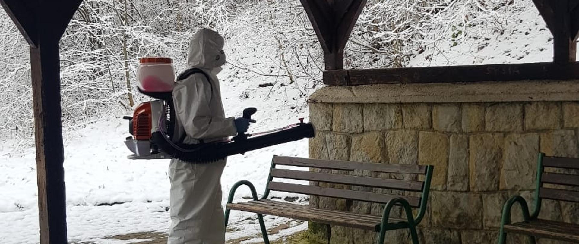 Strażak w kombinezonie dezynfekuje ławkę w miejscu publicznym pod wiatą.