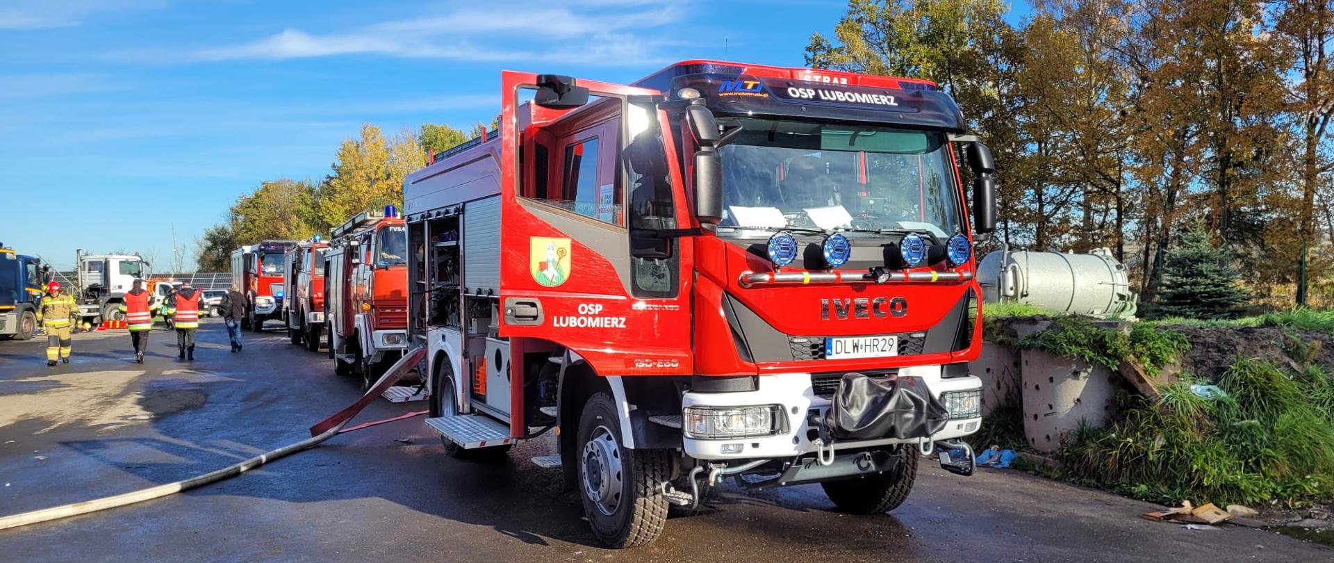 Na zdjęciu widoczne są samochodu ratowniczo-gaśnicze jednostek straży pożarnych, które biorą udział w ćwiczeniach powiatowych. W tle widoczni są ćwiczący strażacy.