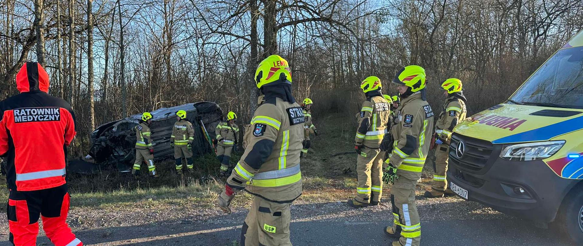 Grupa strażaków i ratownik medyczny na miejscu poważnego wypadku drogowego w lesistej okolicy. Na poboczu leży przewrócony samochód osobowy, wokół którego trwają działania ratownicze.