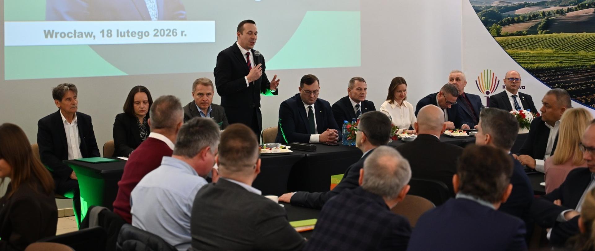 Na sali konferencyjnej na tle wyświetlanej prezentacji, za stołem siedzą dwie kobiety i siedmiu mężczyzn, pomiędzy nimi stoi mężczyzna z mikrofonem i przemawia do osób siedzących przed nim przy stołach konferencyjnych.