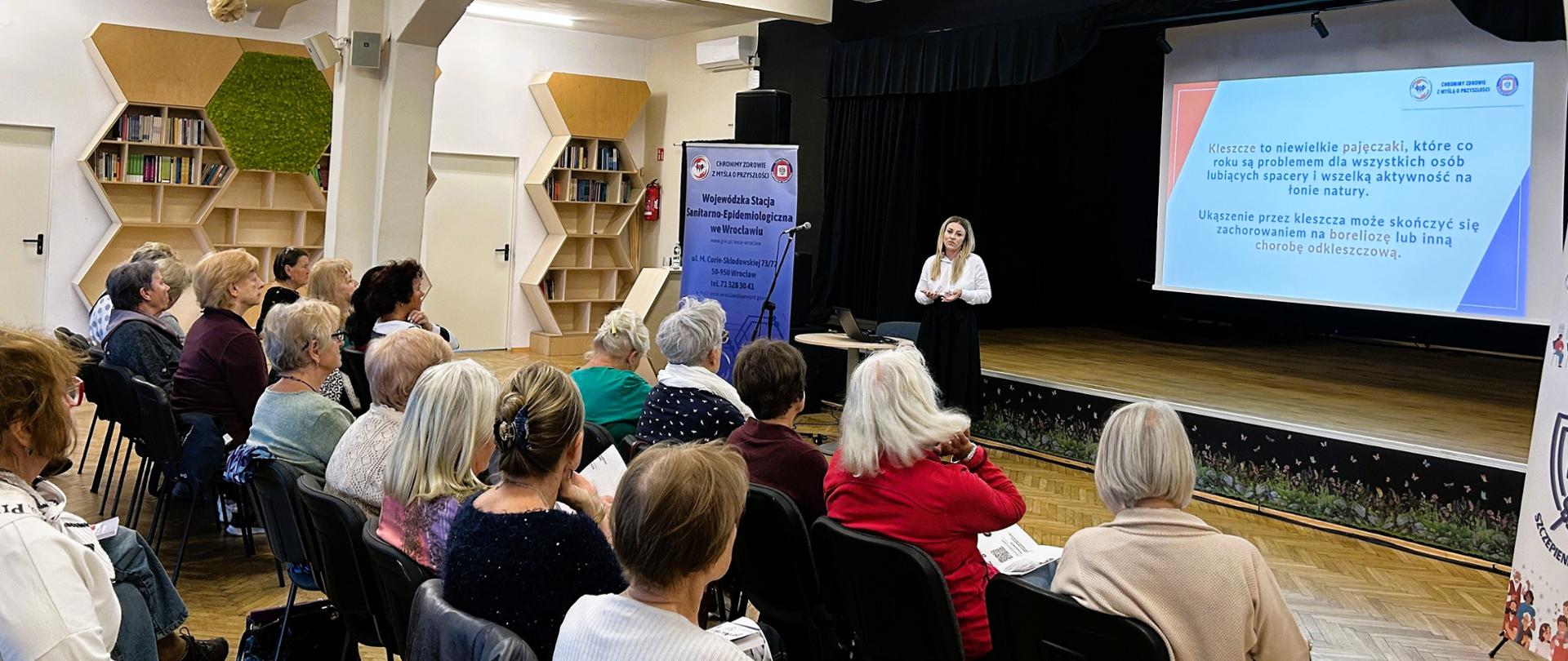 Sala wykładowa wypełniona słuchaczami. Osoba prowadzi prelekcję, za nią na ekranie multimedialnym slajd o zagrożeniu kleszczami