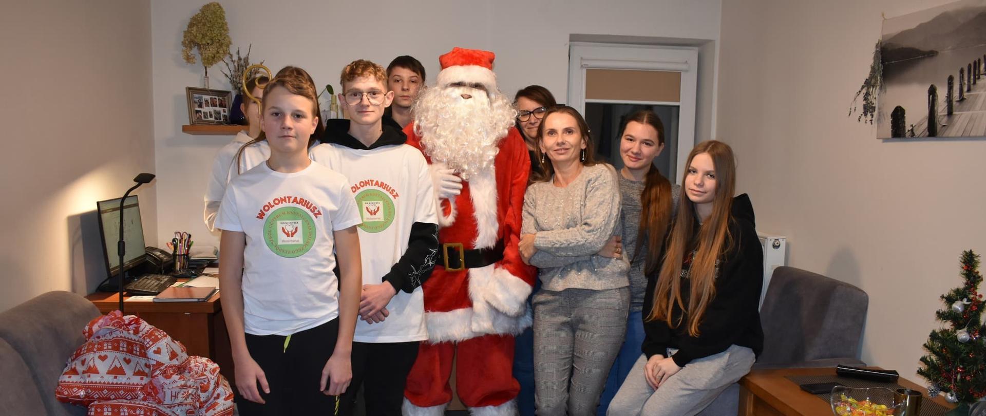 Wspólne zdjęcie wolontariuszy, opiekunów i Świętego Mikołaja w udekorowanym salonie. Wszyscy patrzą w stronę obiektywu, panuje radosna atmosfera.