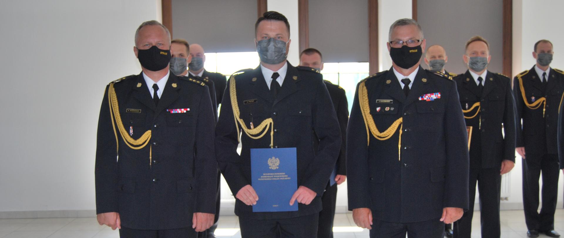 Na pierwszym planie trzech funkcjnariuszy, pozują do zdjęcia pamiątkowego - nowo mianowany naczelnik ośrodka szkolenia w towarzystwie komendanta wojewódzkiego i jego zastępcy. W tle pododdział towarzyszący. Wszyscy w mundurach galowych.