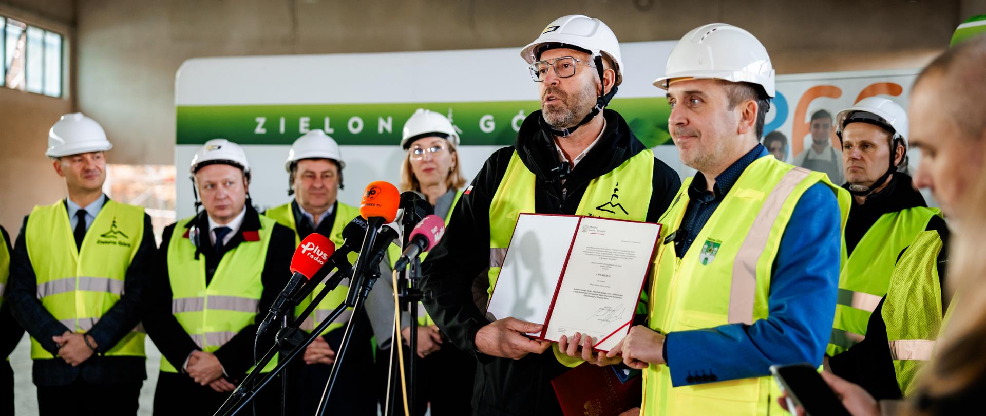 Konferencja prasowa na budowie hali Olimpia. Na pierwszym planie za mikrofonami stoją minister sportu i turystyki Jakub Rutnicki i prezydent Zielonej Góry Marcin Pabierowski, prezentując decyzję o dodatkowym dofinansowaniu. Obaj maja na sobie białe kaski i żółte kamizelki odblaskowe. Za nimi stoją inni uczestnicy konferencji w takich samych strojach.