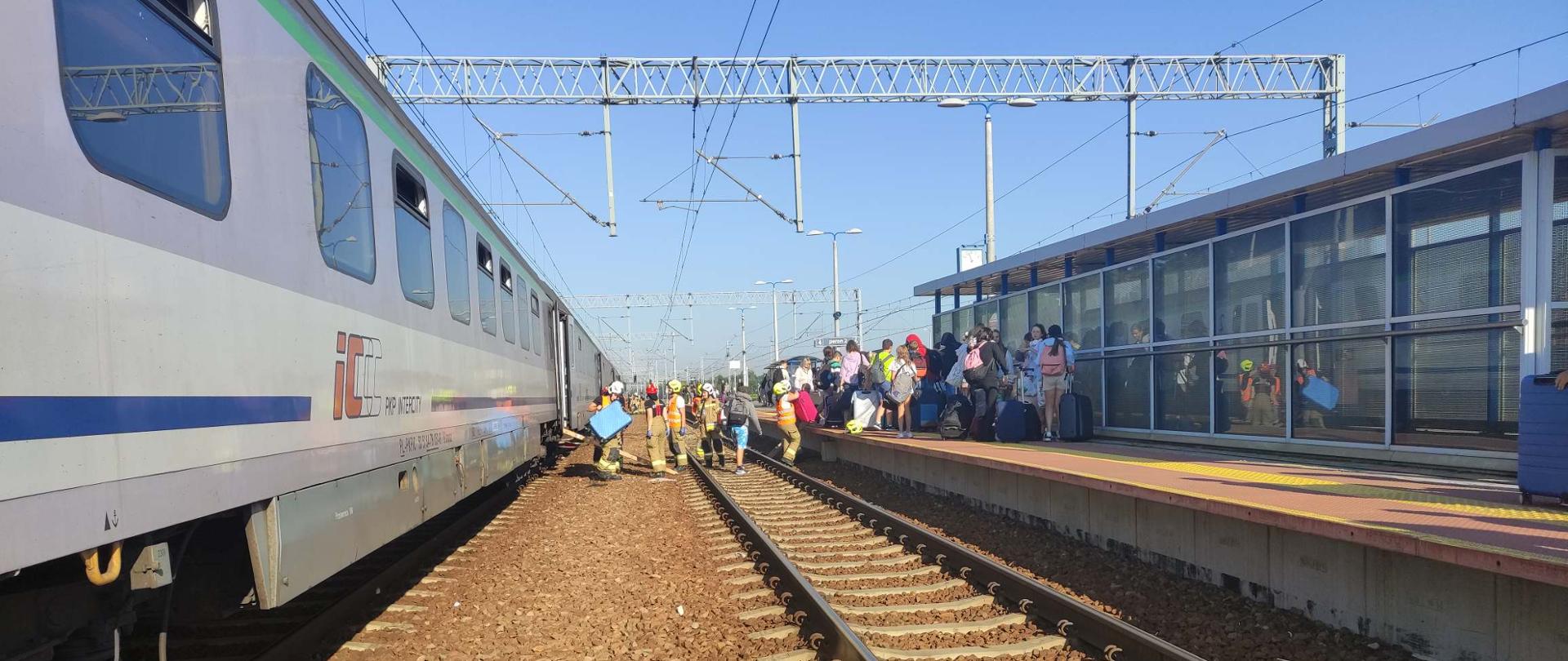 Na zdjęciu wagony pasażerskie pociągu, z którego wysiadają ludzie na sąsiedni peron. Wśród ludzi strażacy w ubraniu specjalnym pomagający w bezpiecznym przejściu.