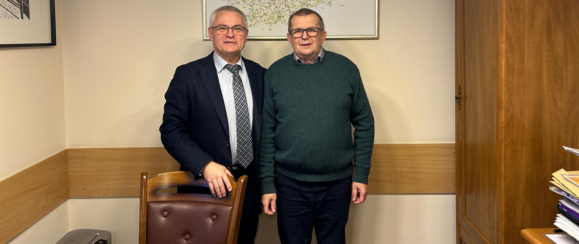 Dwaj mężczyźni stoją w gabinecie urzędowym, pozując do fotografii na tle mapy powiatu elbląskiego