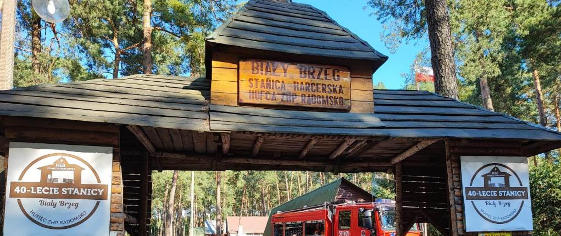 Brama wjazdowa na teren stanicy, w oddali samochód pożarniczy oraz uczestnicy spotkania podczas pogawędki o bezpieczeństwie 