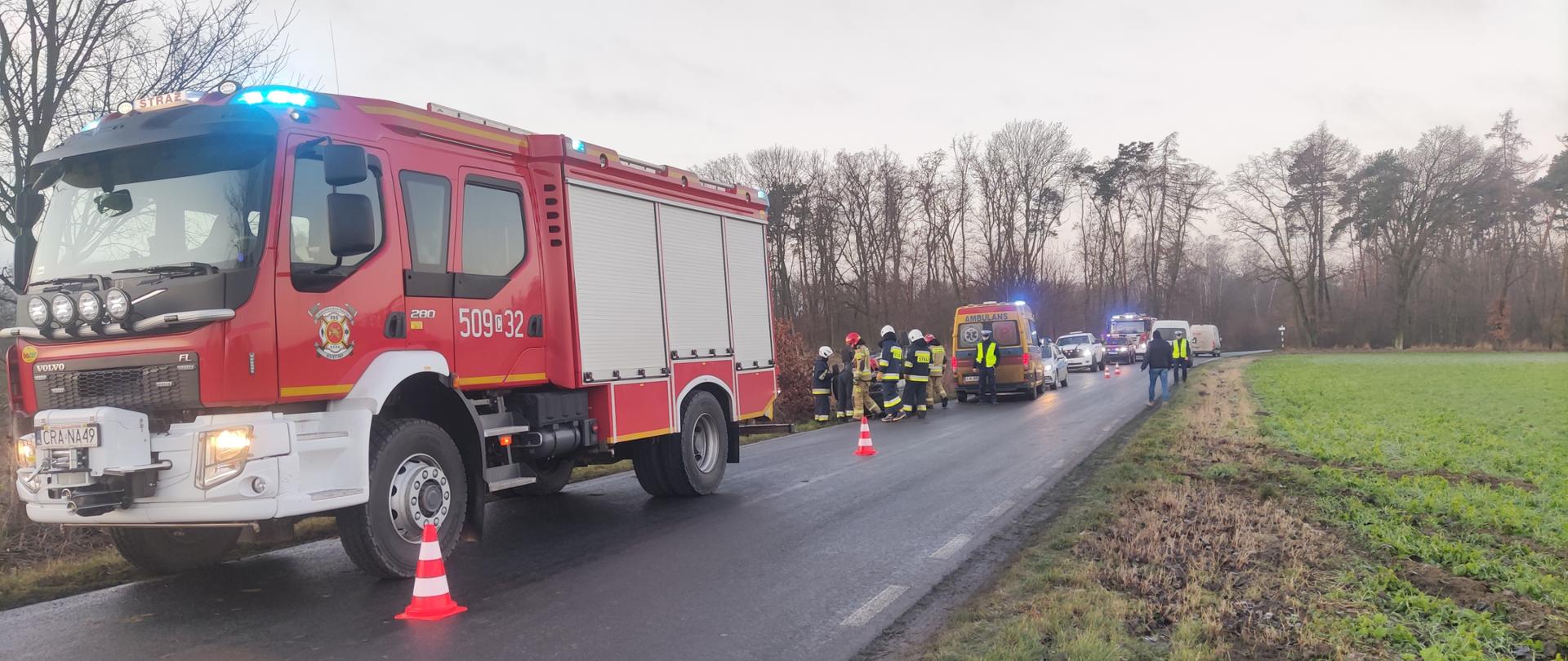 Zdjęcie przedstawia drogę o nawierzchni asfaltowej oraz służby ratownicze podczas kolizji w Żakowicach. Na pierwszym planie wóz strażacki koloru czerwonego, na drugim planie karetka pogotowia, policja i inne pojazdy, strażacy i policjanci podczas czynności służbowych.