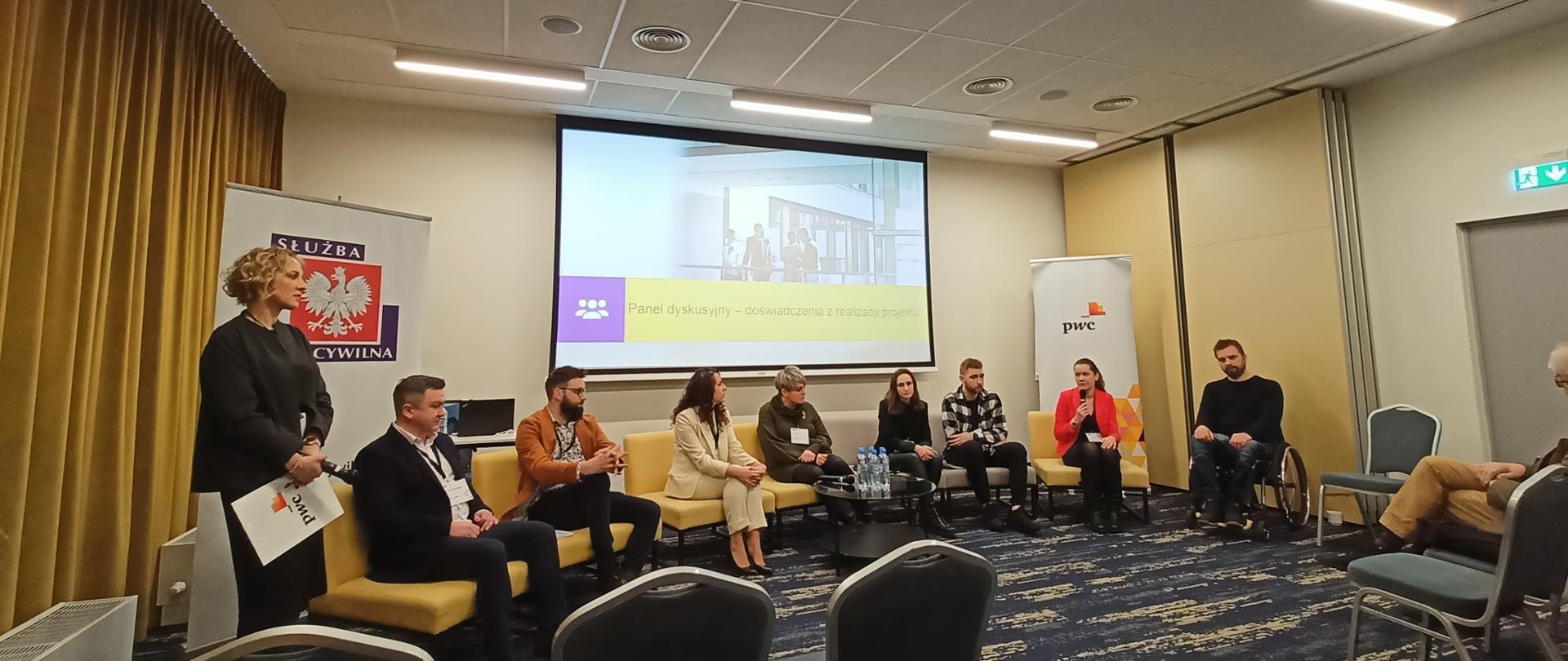 W sali konferencyjnej kilka osób siedzi na żółtych krzesłach, jedna pani trzyma w ręku mikrofon. Jeden z mężczyzn jest na wózku. Na ścianie za panelistami znajduje się duży ekran. 