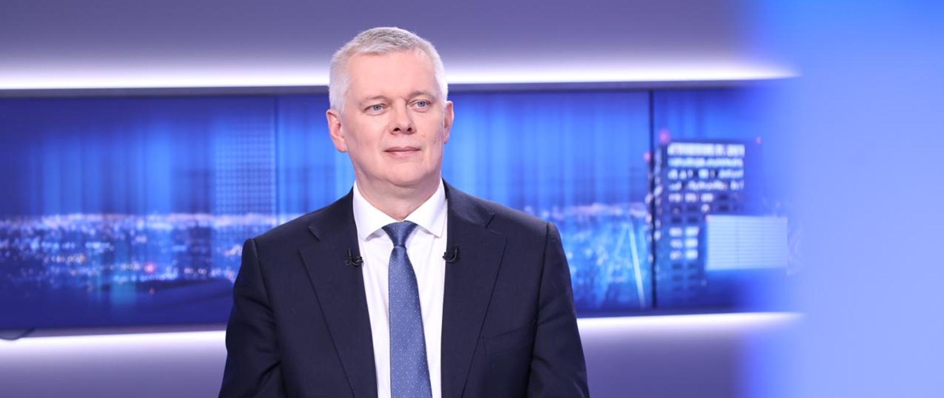 Na pierwszym planie minister Tomasz Siemoniak, siedzący przy białym stole. Na stole znajduje się szklanka z wodą. W tle widać ekran z panoramą oświetlonego miasta nocą.