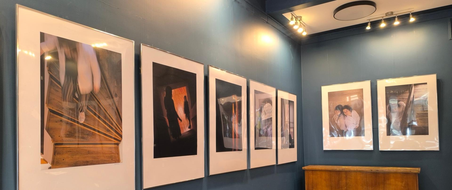 Wnętrze galerii z wystawą fotografii pt. „ONE”. Na ciemnoniebieskich ścianach wiszą oprawione w ramy zdjęcia. W rogu pomieszczenia stoi drewniane pianino z taboretem, a obok fragment niebieskiego siedziska.
