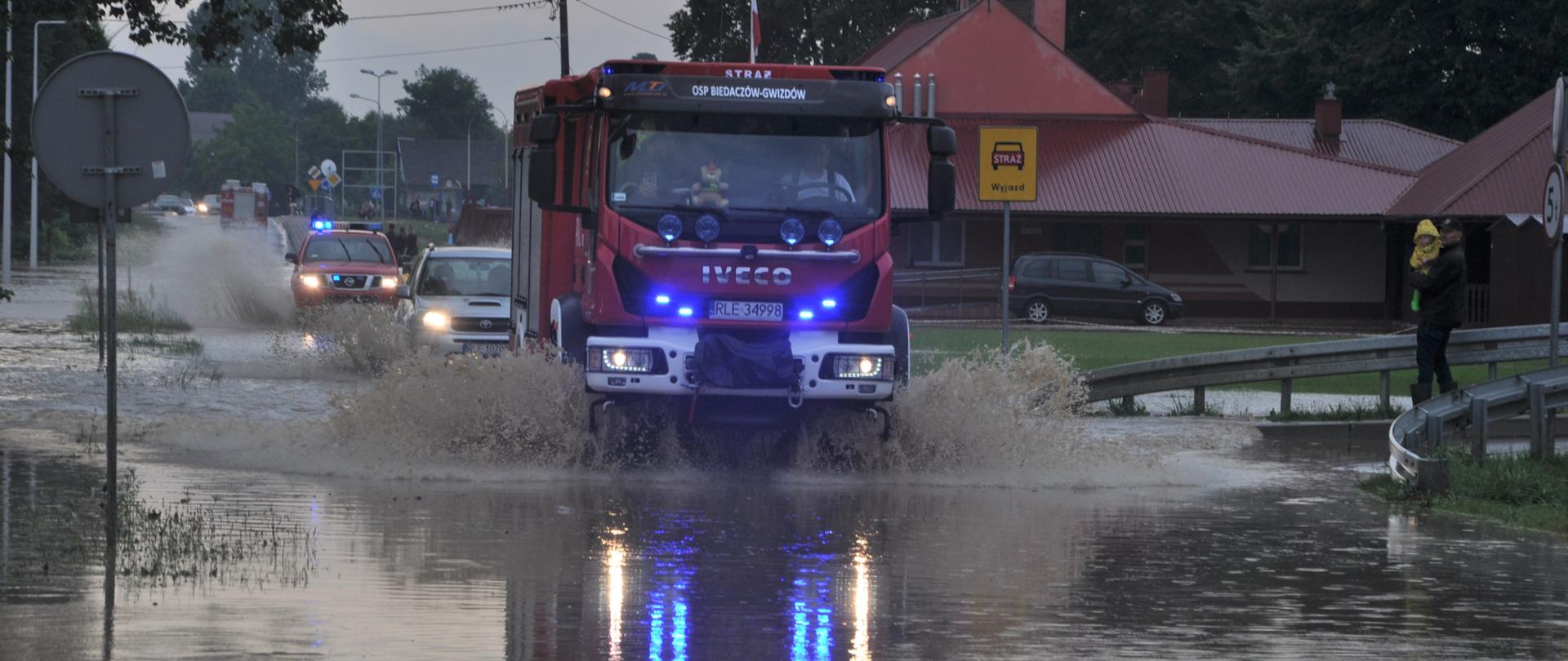 Na zdjęciu widzimy jadące po zalanej drodze samochody pożarnicze.