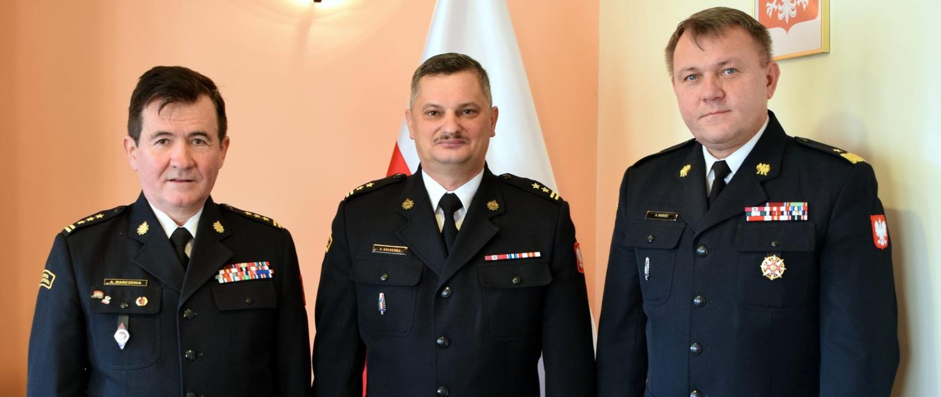 Zdjęcie zrobione wewnątrz pomieszczenia. Na zdjęciu trzech oficerów Państwowej Straży Pożarnej w mundurach wyjściowych.