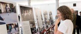 Osoba ogląda wystawę fotograficzną ustawioną w jasnej sali z kolumnami. Ekspozycja składa się z kilku plansz umieszczonych na drewnianych stojakach. Na pierwszej planszy widoczne są fotografie osób grających w piłkę oraz fragment tekstu dotyczącego obozu w Libanie. Kolejna plansza przedstawia stado zwierząt i ma tytuł „Dramat obojętnych przelot bocianów”. W głębi sali stoją pozostali zwiedzający.